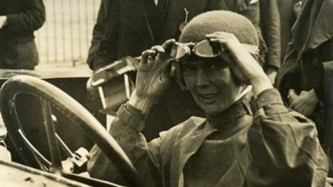 A black and white photo of a woman sitting in a racing car with a large steering wheel. She has a hat and goggles on her forehead and she has both her hands on the goggles. She is smiling and looking at the camera.