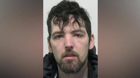 Mugshot of Hepplewhite. He has scruffy black hair and a shore dark beard.