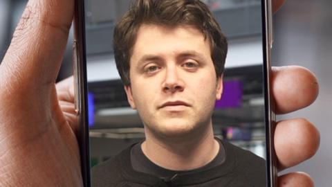 A man with brunette hair is looking towards the camera and is in a phone held by a hand.