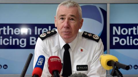 A man sits at a table with several mics belonging to media organisations in front of him. Lapels are on his shirt. The word police is printed on his shirt on a badge. 