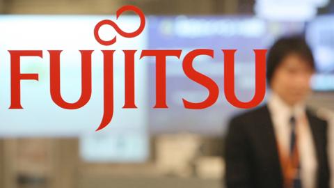 Fujitsu logo is seen pictured on glass with an out-of-focus employee in the background, taken at the company's offices in Tokyo in 2007