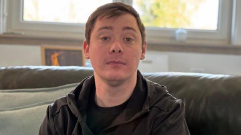 Stephen Reside, who has brown hair combed in a side shed, sits on a leather couch in a living room. he is looking directly at the camera with a serious expression on his face.
