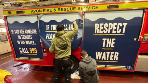 Two men apply posters to the side of a fire engine