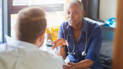 A GP and patient chat inside a treatment room. The GP is facing the patient who cannot be fully seen. The female GP has cropped grey hair and a blue shirt.