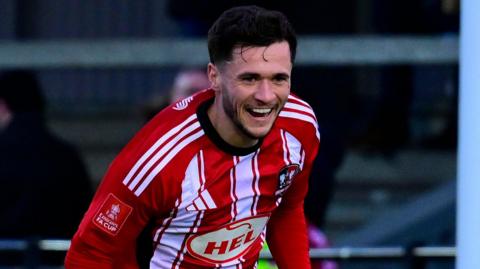 Jack Aitchison with short black hair and a neat beard is smiling. He is wearing th Exeter City home strip with red and white stripes.