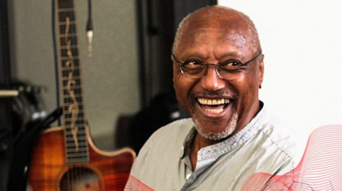 A man with glasses, a faint white beard and a white top with a wood-paneled guitar behind him