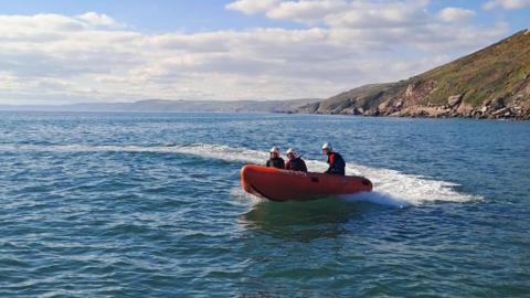  A picture of lifeguards on a lifeboat travelling across a stretch of water. The boat is orange in colour and there is three people sat on it. There is a large rock face in the distance.