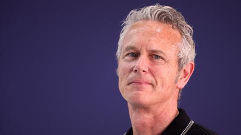 Mark Foster stands against a deep purple background wearing a black polo shirt. He is smiling and looking directly at the camera