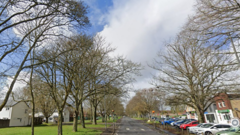 A view of the main road through Larkhill from Google, with homes, grass and trees on one side and shops and cars parked on the other.