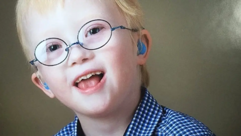A portrait picture of James Dwerryhouse. He has blond hair and is wearing glasses with a round frame. His mouth is open and he is looking at the camera. He is wearing a blue chequered shirt.