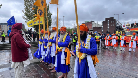 Five men wearing blue robes with orange and white. They are all holding yellow flags and other members of the procession and crowd can be seen behind them