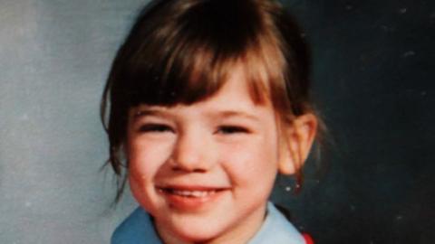 A young girl with brown hair in a red school jumper smiles at the camera