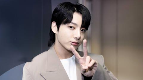 Jung Kook in a beige suit with white t-shirt underneath. He's in a studio setting. He's smiling at the camera and doing a peace sign - his index and middle fingers extended. His hair's styled in curtains, and he has a lip piercing.