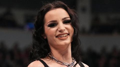 Saraya smiles at the camera. She's in the middle of a wrestling ring, holding a microphone and wearing a vest top and chunky chain necklace