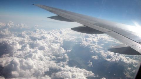 The view from above the clouds from the left side of an unidentified airplane, with the left wing visible in the foreground and white clouds separating to show the mountainous ground below