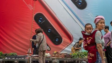 A woman carries a child pasased a billboard advertising smartphones for China's Huawei Technologies Co., at a market on June 1, 2019 in Mangshi, Yunnan Province, southwestern China.