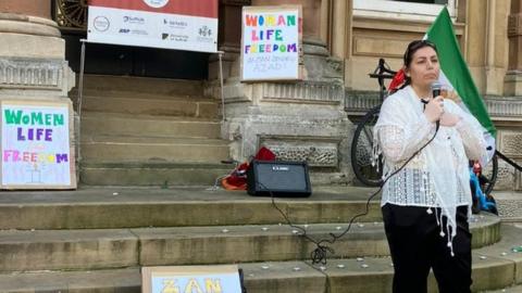 A woman speaks at a vigil for Iran held in Ipswich. There are signs behind her that read women, Life, Freedom.