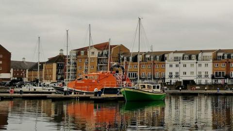 An orange boat sits in the centre of the harbour with houses and grey sky behind and water in front