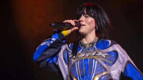 Billie Eilish performing on stage in August 2023. Billie is a 22-year-old white woman with long black hair worn loose. She has a fringe to her eyebrows and wears a graphic long-sleeved blue and grey top. She holds a microphone up as she sings.