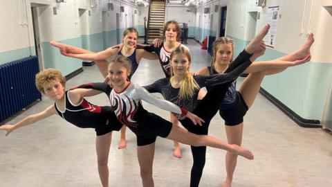Six young gymnasts holding their legs up in a prison corridor
