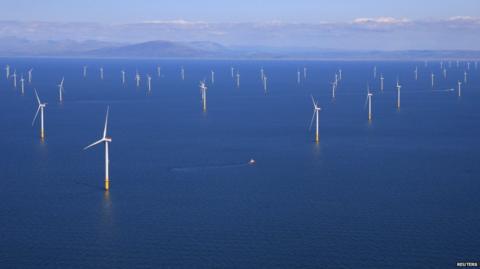 Dozens of wind turbines are shown across a deep blue ocean with a land mass in the background