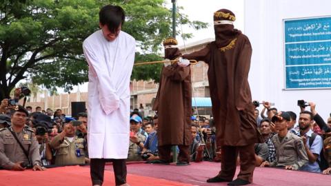 Man in white gown getting caned in front of a crowd