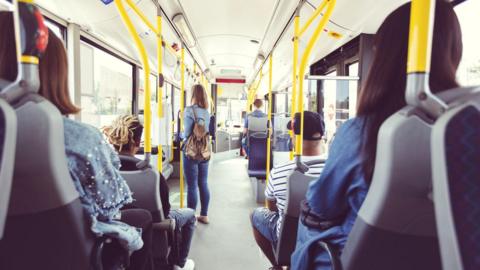 A shot from the back of a bus. It shows five people sitting on rows on either side with one woman standing and holding on to a pole. She is wearing jeans, a denim jacket and has a brown backpack slung over her left shoulder.