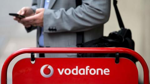 A man uses a mobile phone next to a Vodafone sign