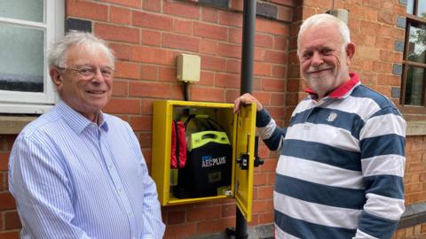 David Dey in a blue and white striped shirt, standing with Howard Painter, in a navy and blue striped shirt. Between them is a wall-mounted yellow defibrillator box that is open. Inside is a black defibrillator box reading "AED PLUS", next to that is a red bleed kit 