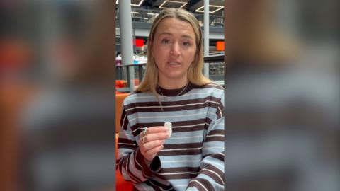 Lucy Woodham looks at the camera while holding a small white mic. She is wearing a blue and brown striped jumper.