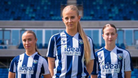 Three female football players stand in blue and white West Bromwich Albion kits in a football stadium.