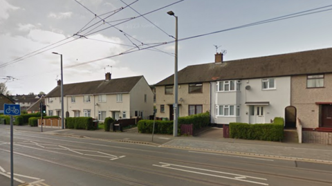 A view of a street with tramlines overhead and a row of semi-detached and terraced houses.