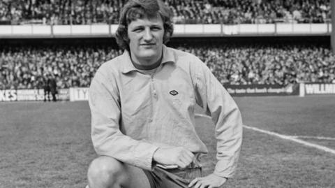 Jimmy Bone in a black and white photo before a game for Norwich City at QPR in April 1972, His left knee is on the pitch, his right knee up and his foot on the ground. His right elbow is posed on his thigh. He is wearing a long-sleeved top, and a packed terrace can be seen behind him 