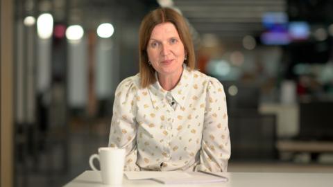 Bethan Lewis, a woman with shoulder length light brown hair and a floral shirt adresess the camera.