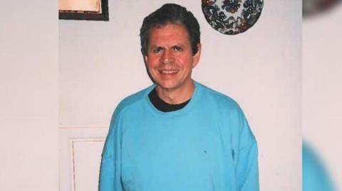 Man wearing blue jumper and smiling inside his home. 