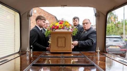 A funeral director team putting a coffin with flowers laid on top into a hearse. They are in a residential area and are all wearing suits. The view is from inside the hearse