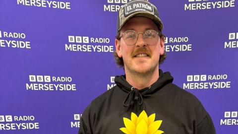 Joe Jones is wearing a baseball hat and a black sweatshirt with a bright yellow glowing flower and is smiling. He is standing in front of a purple display with BBC Radio Merseyside written on it.