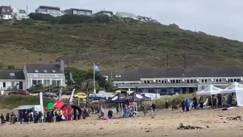 A group of surfers and supporters are gathered at the top of the sandy beach. There are branded pop-up banners and gazebos. Behind the beach are houses. 