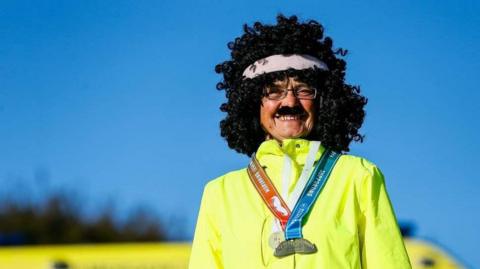 A person in a high-visibility yellow jacket wearing a MoRun medal with red and blue ribbons, a fake moustache and a wig with a white headband.