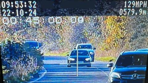 The view from a speed camera. A dark car is in the middle of the road, as if overtaking. A white cross, like an addition sign, is covering the car. 