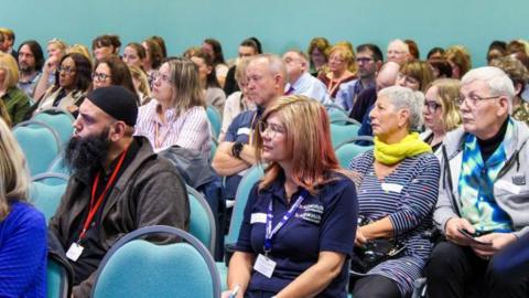 A group of people are sitting in an audience, on blue chairs, one woman has a shirt with "Healthwatch Cambridgeshire" written on it