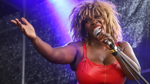 Sass is in a red leather dress and wig to look like Tina Turner. She is singing and has a microphone in her hand close to her face. 