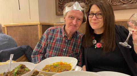 Tim Hartley and his wife Helen Lucitt are sitting at a table full of different curry dishes.  Tim is sat on the left of Helen wearing a chequered blue and red shirt. He is wearing a silver paper crown from the crackers. Helen is lifting up a glass of white wine. She is wearing glasses and a black top.