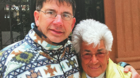 Adrian has glasses on with dark coloured hair and is wearing a patterned fleece jacket. He has his arm around his mother Rita who also wears glasses and is wearing a yellow jacket