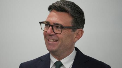 Andy Burnham looks off camera to the left. He is wearing a dark suit, white shirt and dark tie.