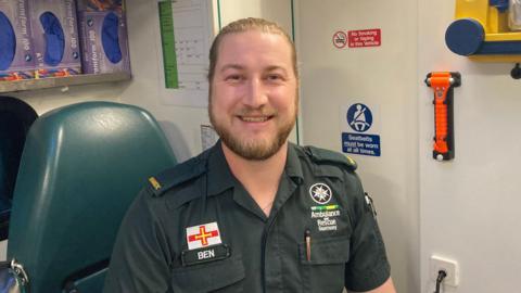 Ben Adam sat in a green chair. He has his name 'Ben' written on his left with a flag of Guernsey above it. On the right is 'Ambulance and Rescue Guernsey'. 