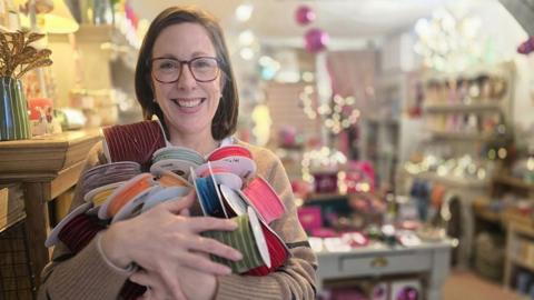 A woman with dark shoulder length hair and glasses on stands with lots of spools of ribbon in her arms. Behind her is a blurred background showing a shop with tables and cabinets with items on them and fairy lights dotted around.