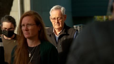 Ian Wilkinson wearing a black vest and spectacles, with a head of white hair. He is walking, surrounded by several others.