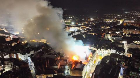 A drone shot of smoke coming from a building's roof which is on fire. There are two aerial platforms above the fire.