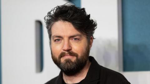A man with dark hair and a dark wearing wearing a black shirt.